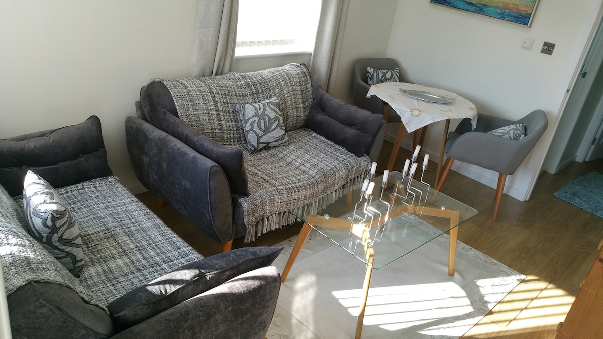 A comfortable seating area is furnished with two dark gray sofas, each adorned with decorative pillows. A glass coffee table sits in the center, complemented by four modern dining chairs next to a small table. Natural light filters through a window with sheer curtains.