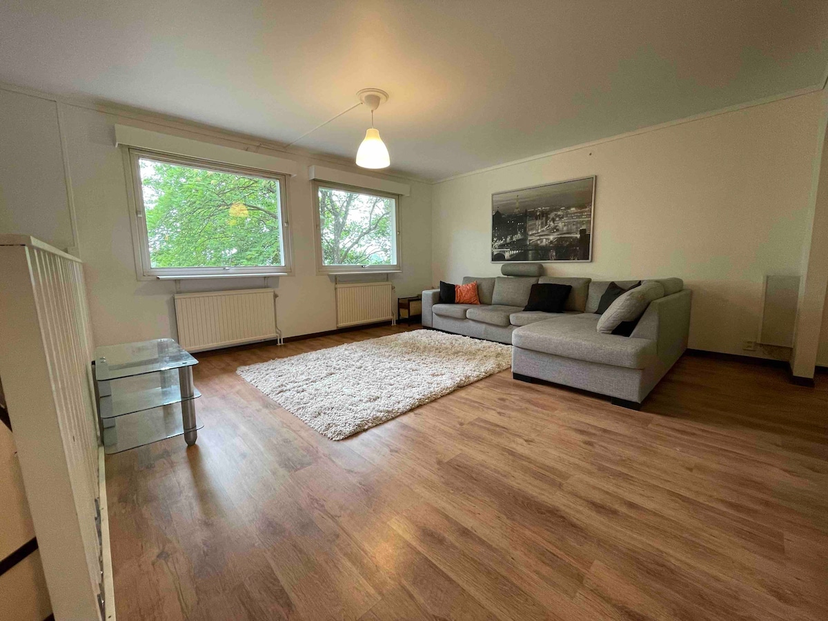A spacious living area features a comfortable gray sectional sofa complemented by a decorative pillow. A soft area rug lies on the wooden floor, and a glass coffee table is positioned nearby. Natural light enters through large windows adorned with simple treatments.
