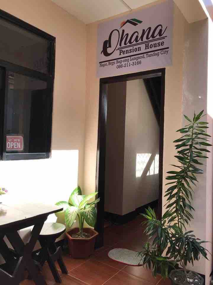 The entrance to Ohana Pension House is visible, featuring a welcoming sign above the door. A small table with chairs is placed outside, alongside a potted plant. Natural light is captured through a window, illuminating the space.