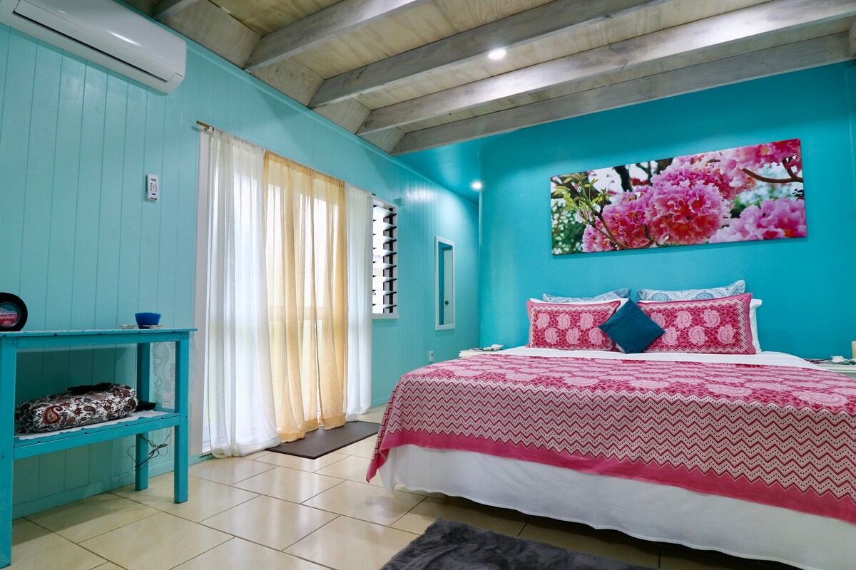A spacious bedroom showcases vibrant turquoise walls and a colorful floral artwork above the bed. The large bed is adorned with decorative pillows, while natural light streams through white curtains. An air conditioning unit is mounted on the ceiling, ensuring comfort.