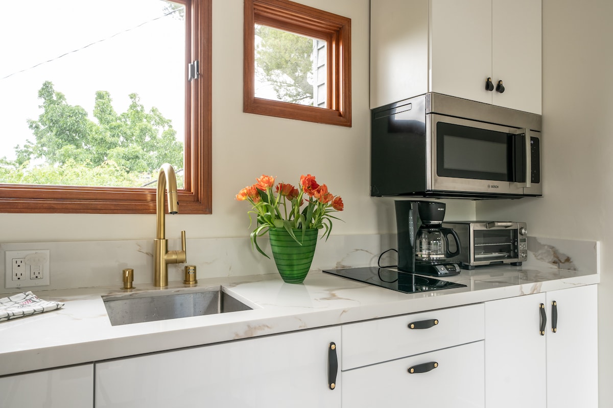 A modern kitchen space features white cabinetry and a marble countertop. A stainless steel microwave is positioned above a countertop oven. A coffee maker and an attractive vase with flowers are placed on the counter, while a window allows natural light to illuminate the room.