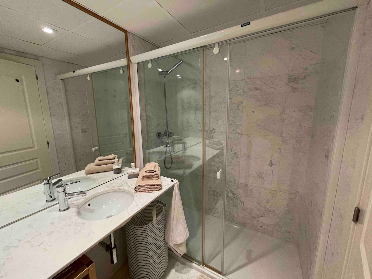 A modern bathroom is presented, featuring a spacious glass shower enclosure with a rainfall showerhead. A double sink vanity is visible, adorned with neatly arranged towels in natural tones. Marble walls add a touch of elegance to the overall design.