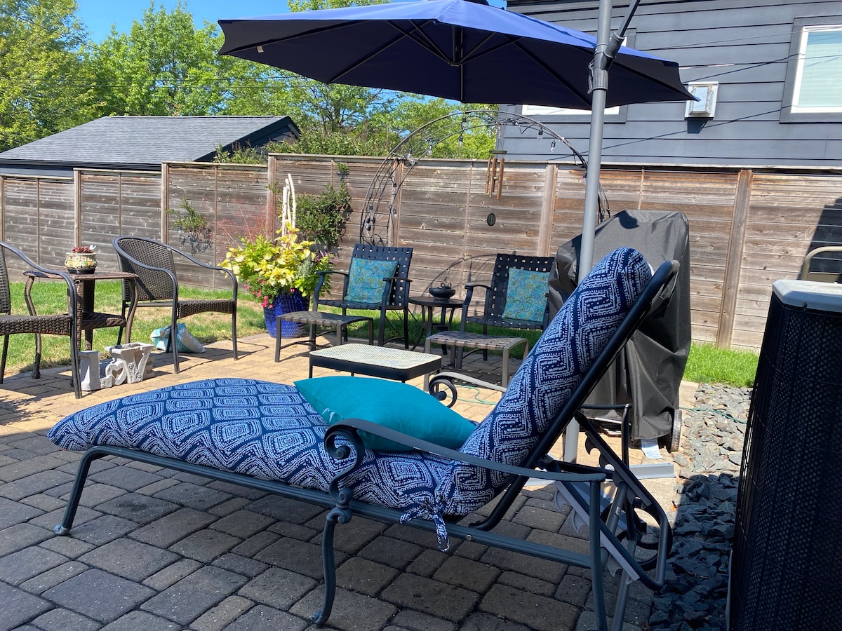 An outdoor patio area is visible, featuring a patterned lounge chair beneath a blue umbrella. A small table accompanies the chair, surrounded by additional seating options. Lush green landscaping and a wooden fence provide a sense of privacy and tranquility.