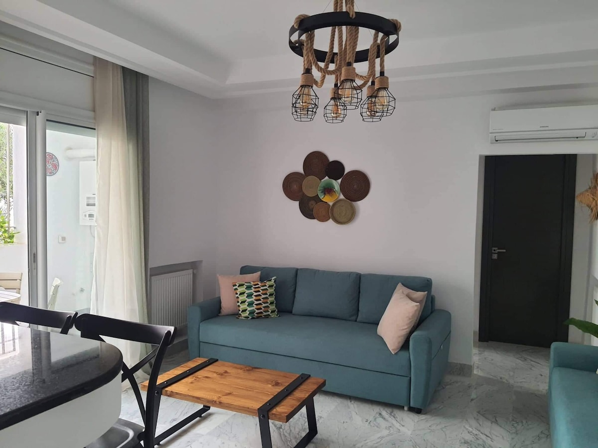 A modern living area features a light blue sofa adorned with two pillows and positioned near a wooden coffee table. A decorative wall installation made of woven materials adds texture, while natural light illuminates the space through glass doors leading to a terrace.