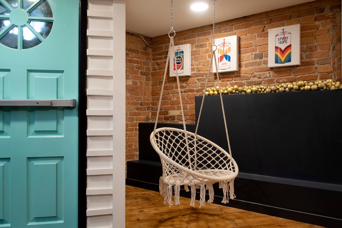 A cozy entryway is seen with a vibrant teal door and exposed brick walls. A macramé swing chair hangs gracefully, positioned in front of an artistic setup featuring framed prints. Warm wooden flooring adds to the welcoming ambiance.