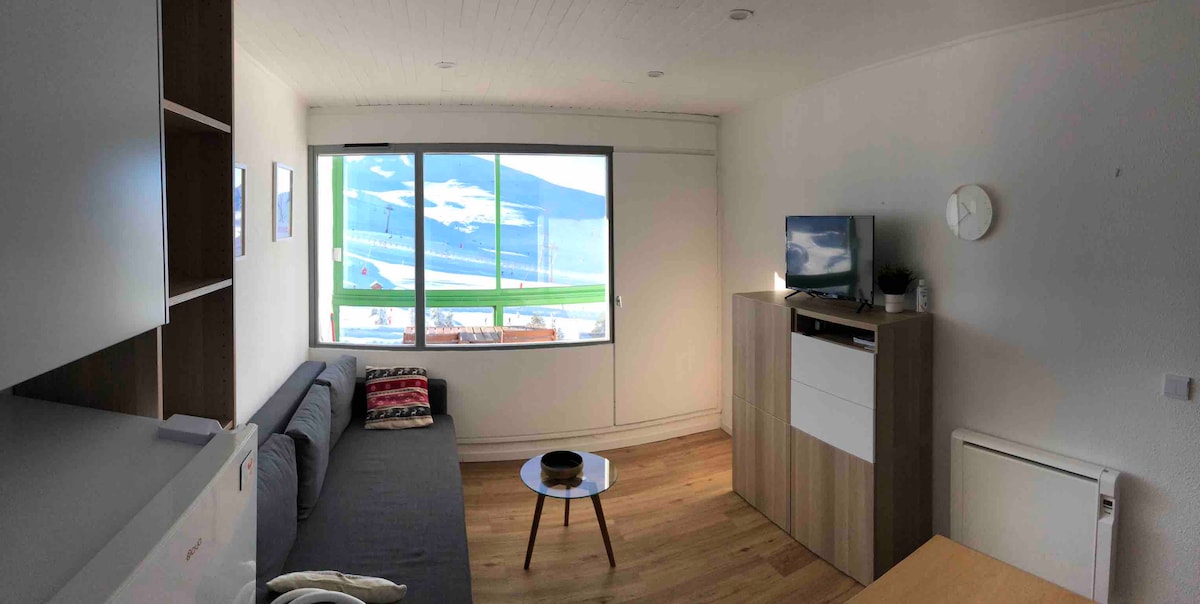 A bright living area is furnished with a comfortable gray sofa and a small coffee table. A television is mounted on the wall. Large windows provide a panoramic view of the scenery outside, enhancing the room's natural light.