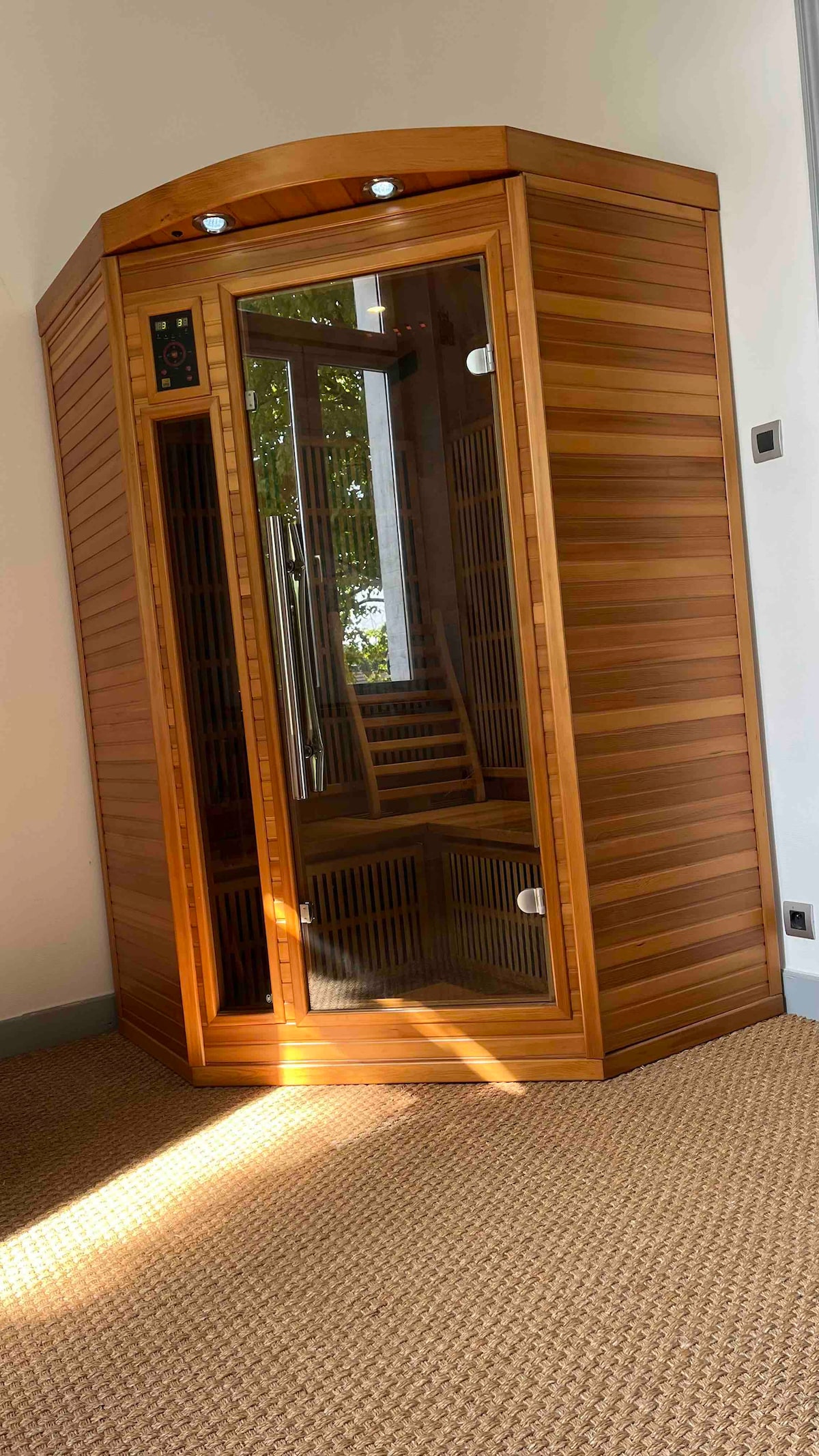 A corner sauna is set against a neutral wall, showcasing a wooden exterior with glass panels. Soft light filters through, highlighting the interior's seating. The warm tones of the wood create a serene environment, complemented by a textured carpet beneath.
