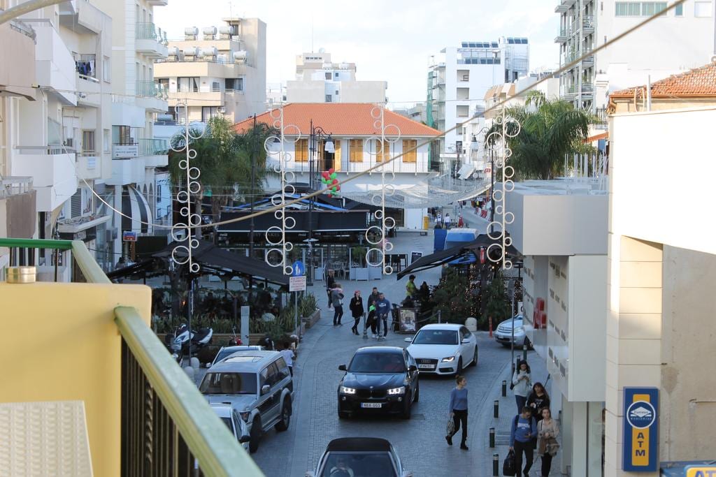 An outdoor view from a balcony showcases a vibrant street scene lined with buildings. Pedestrians and vehicles can be seen moving along the bustling road, which features palm trees and festive decorations. A variety of cafes and shops are visible, enhancing the lively atmosphere.