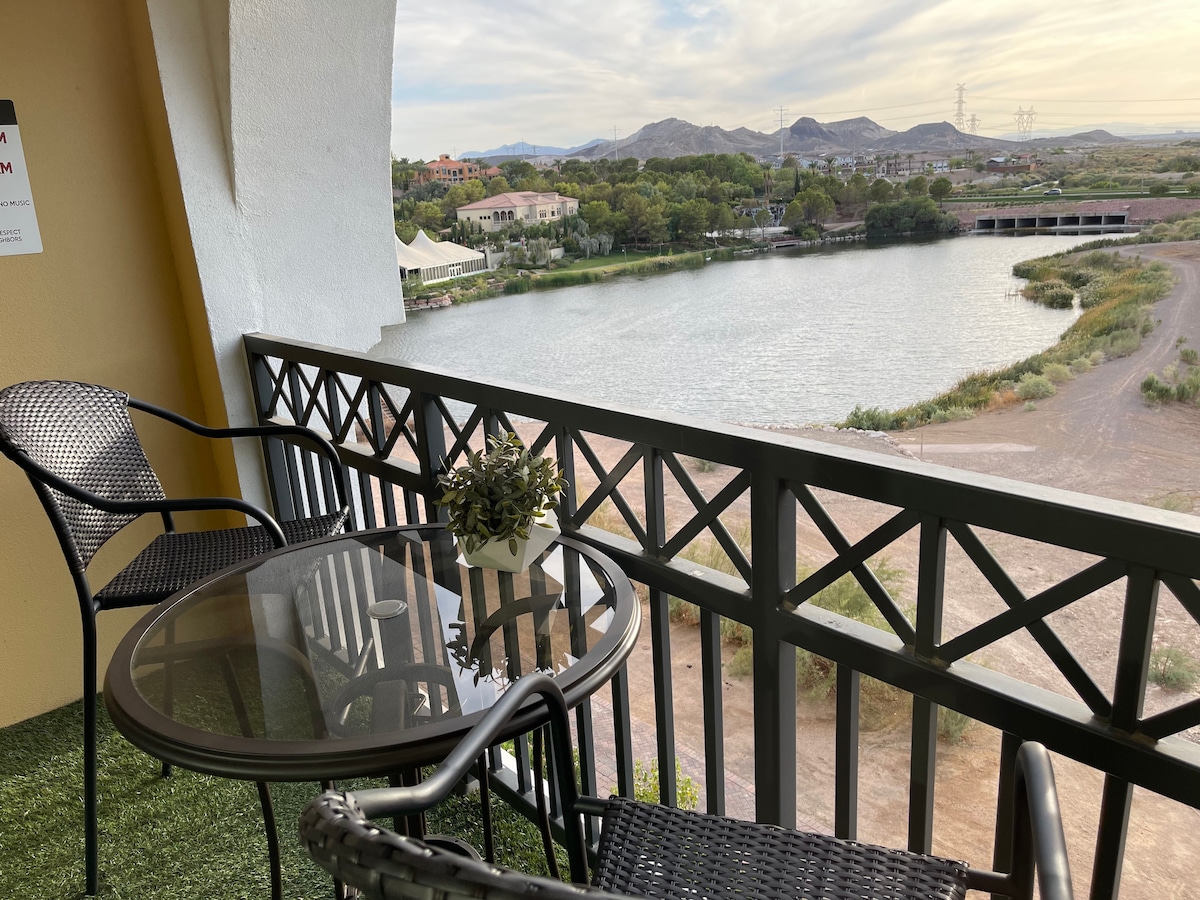 A private patio features a glass-top table surrounded by two black chairs. Lush greenery decorates the table, and the railing offers views of a serene lake and distant mountains under a partly cloudy sky.