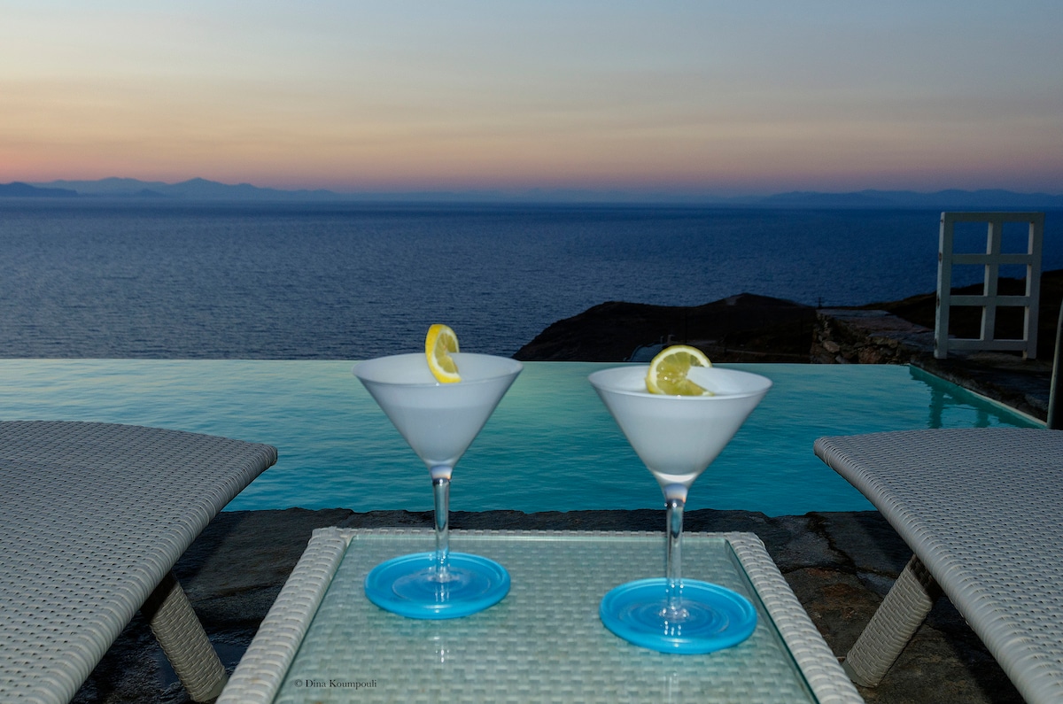 Two martini glasses with lemon slices are placed on a glass-topped table, overlooking an infinity pool that merges with the serene sea at sunset. The calming colors of the sky transition smoothly from soft pastels to deeper hues, enhancing the tranquil atmosphere.