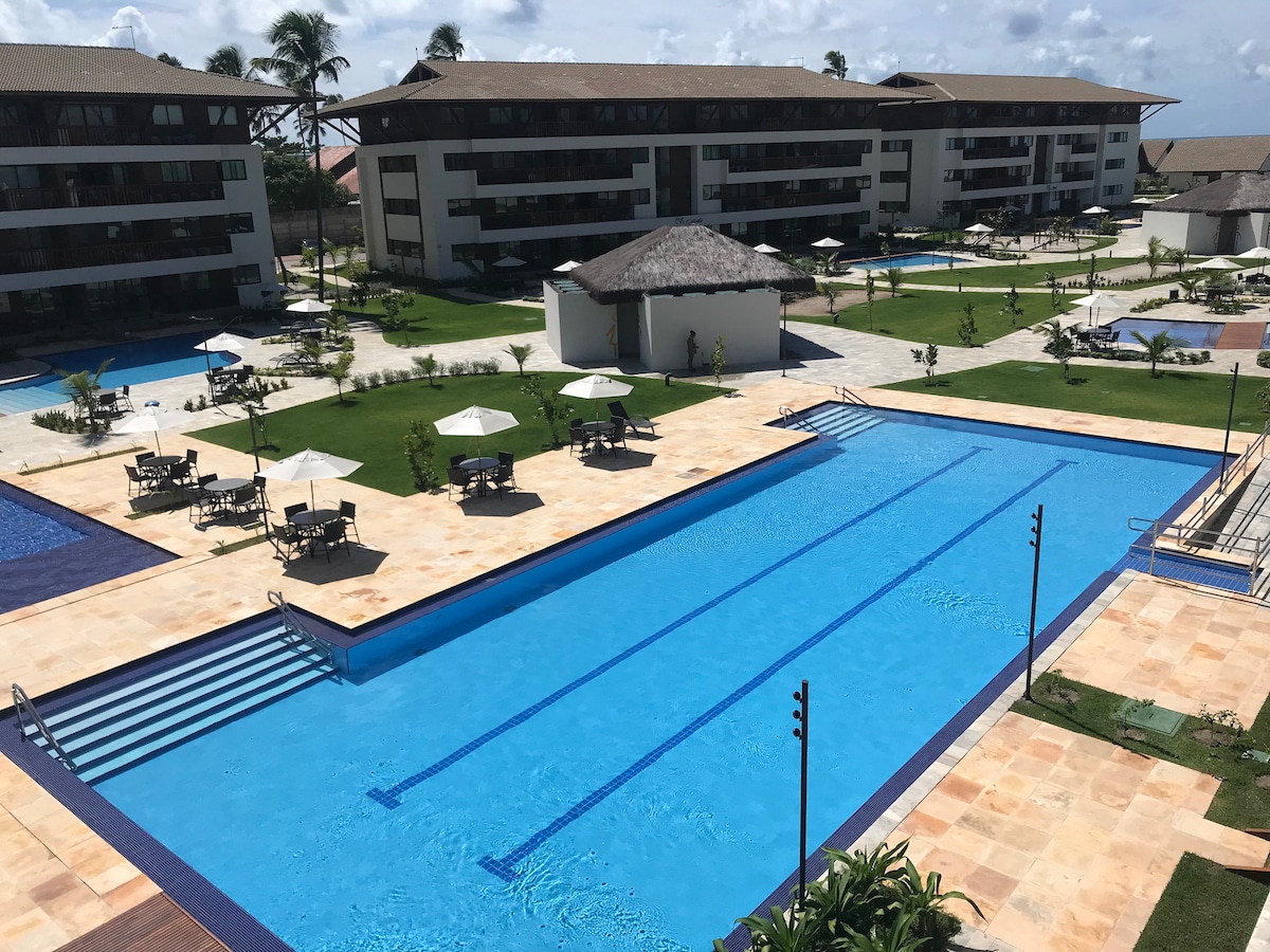 A large swimming pool is centrally located, surrounded by manicured greenery. Sun loungers and umbrellas are positioned nearby for relaxation. The scene includes multiple low-rise buildings with balconies and a thatched-roof structure, all set against a backdrop of partial clouds and blue sky.