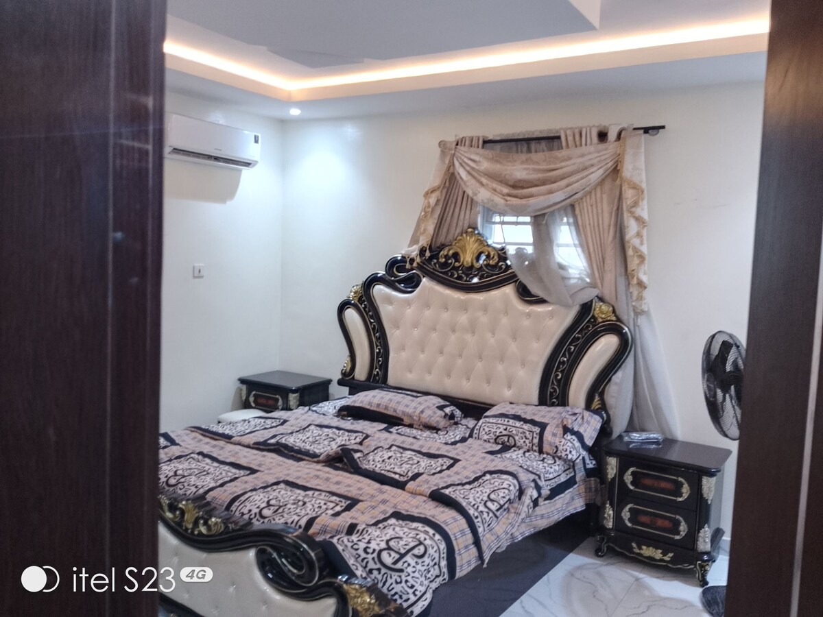 A spacious bedroom features an ornate bed with a tufted headboard and intricate detailing. Soft lighting highlights the room's elegant decor. Air conditioning is present, and a fan stands nearby, ensuring comfort. A window with flowing drapes allows natural light to filter in.