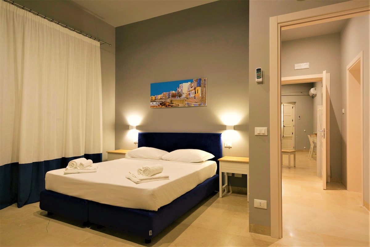 A spacious room is featured with a large bed adorned with neatly arranged linens. Soft lighting is provided by bedside lamps. A large window with curtains allows natural light to brighten the space. An artwork depicting a scenic view is hung above the bed.