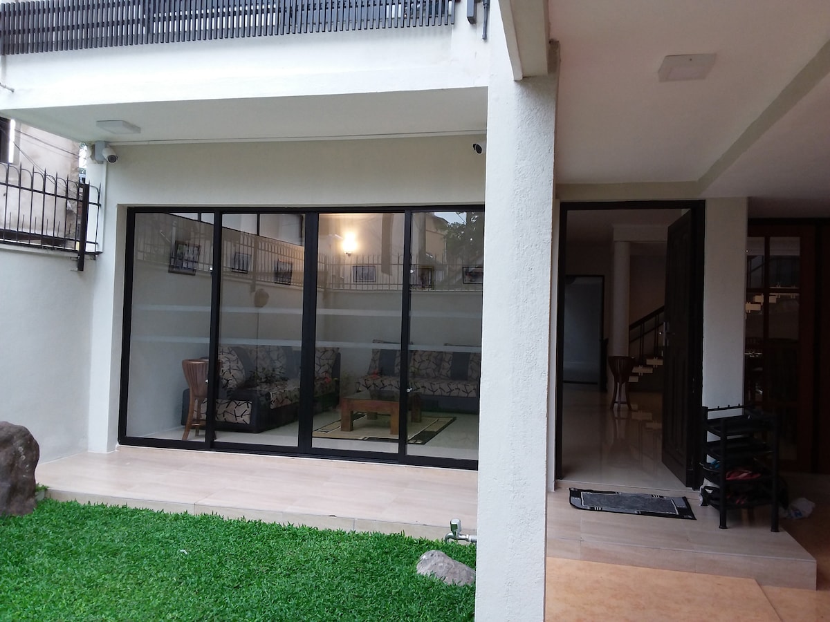A well-lit outdoor seating area features large glass sliding doors that provide a view into the living space. Comfortable seating is arranged on a patterned rug, complemented by a small table. A manicured lawn surrounds the space, enhancing the inviting atmosphere.