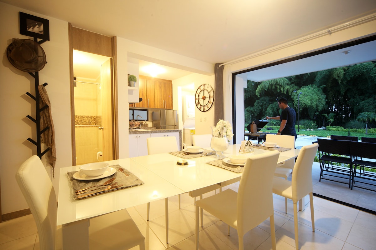 A modern dining area is set with a white table surrounded by six chairs. A view of a lush green outdoor space is visible through large sliding glass doors. The adjacent kitchen features wooden cabinetry, enhancing the open layout.