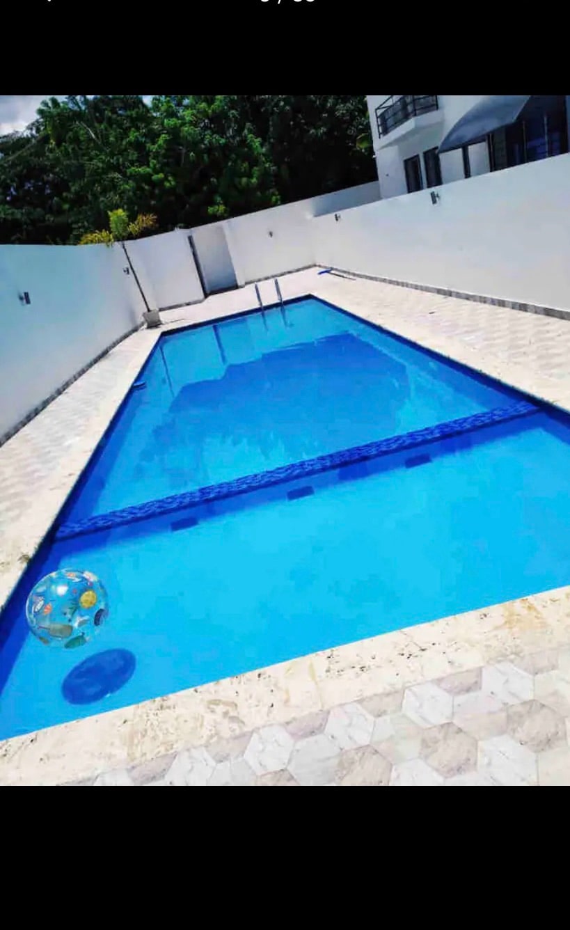 An outdoor swimming pool is displayed, featuring clear blue water and surrounded by white walls. The pool is positioned with steps leading in, and the surrounding area is paved with patterned stone tiles. A beach ball floats gently on the water's surface.