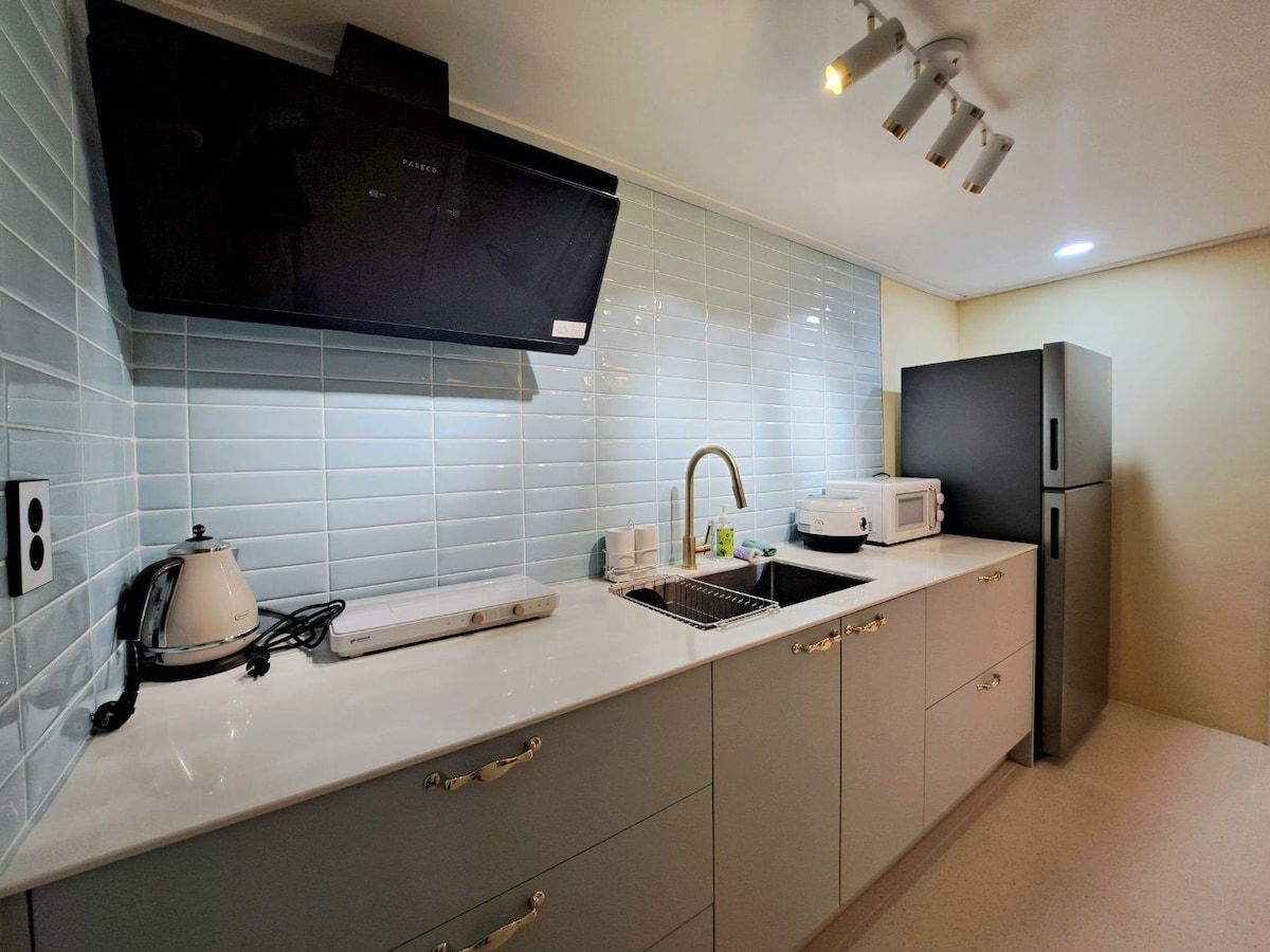 The kitchen features modern cabinetry in a soft grey finish and a white countertop. A stainless steel sink is paired with a sleek faucet. Appliances include a microwave, kettle, and refrigerator. The backsplash consists of light blue tiles, enhancing the fresh atmosphere.