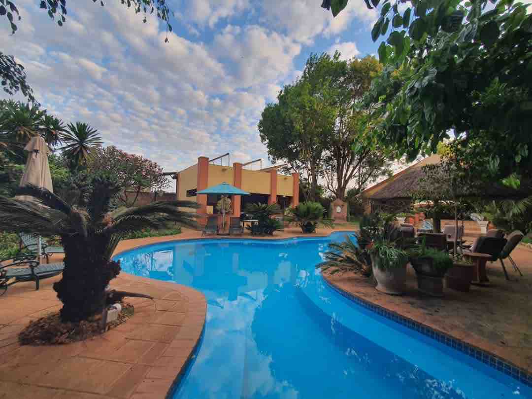 A clear blue swimming pool is surrounded by lush greenery and tropical plants. Comfortable seating areas are situated under umbrellas, with a thatched roof structure slightly visible in the background. Soft clouds drift across the sky, enhancing the peaceful environment.