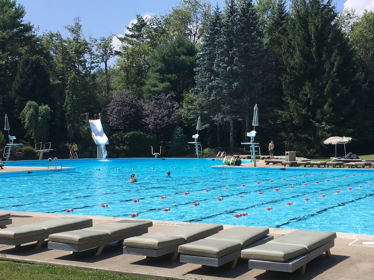 A spacious community pool is surrounded by lush greenery and tall trees. Several lounge chairs in muted colors are positioned beside the water. A water slide and diving boards are visible at one end, with swimmers enjoying the clear, inviting water.