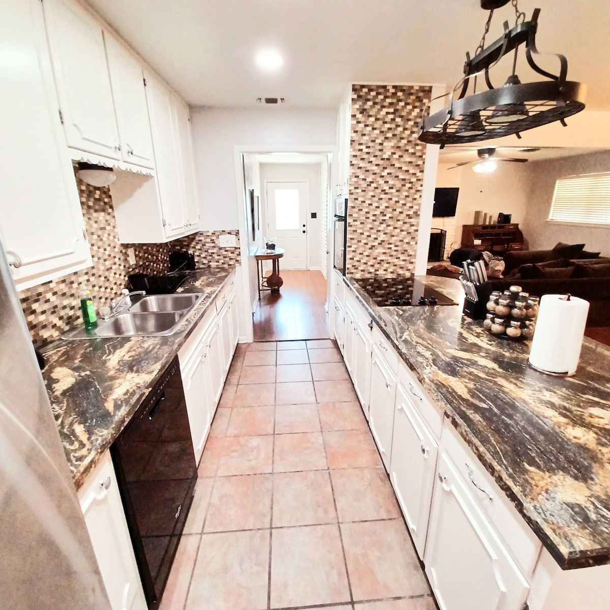 The kitchen features dark granite countertops paired with white cabinetry, providing ample workspace. A stainless-steel refrigerator and black appliances are visible. Neutral tile flooring extends throughout, while a decorative backsplash adds texture. A door leads to the adjacent hallway, enhancing the open feel of the space.