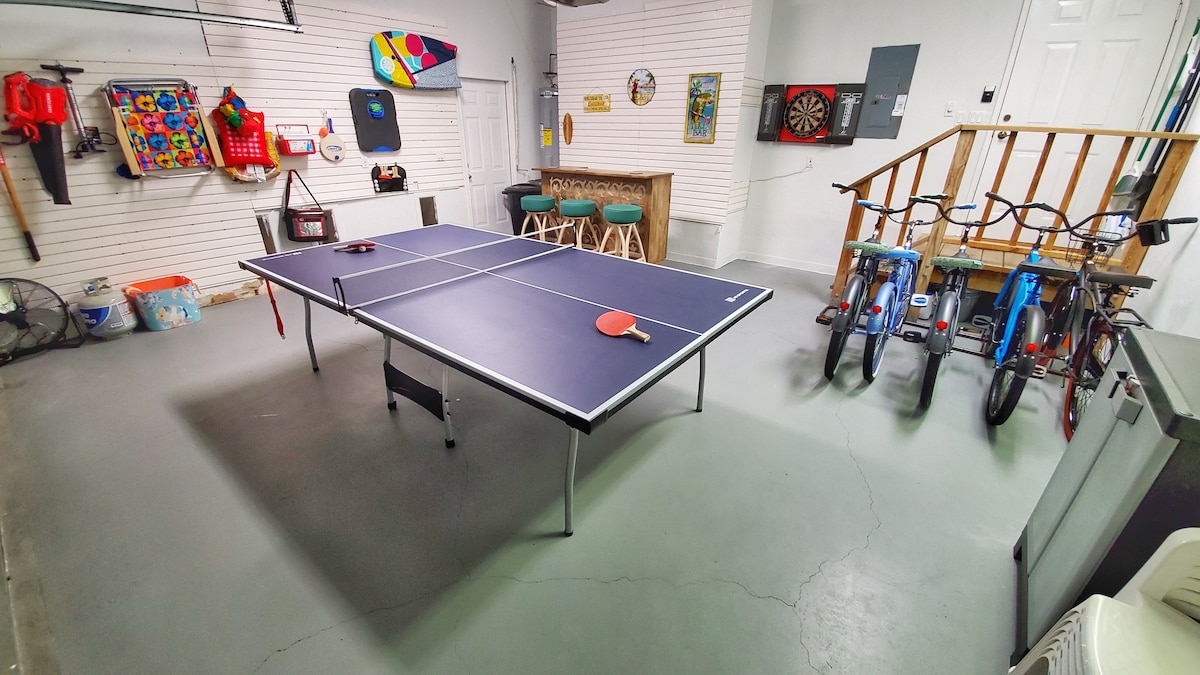 The spacious game room features a blue table tennis table centrally located, with a ping pong paddle resting on it. Bicycles are parked against the wall, and colorful games and decor adorn the surrounding space. A bar-style table is positioned in the background.