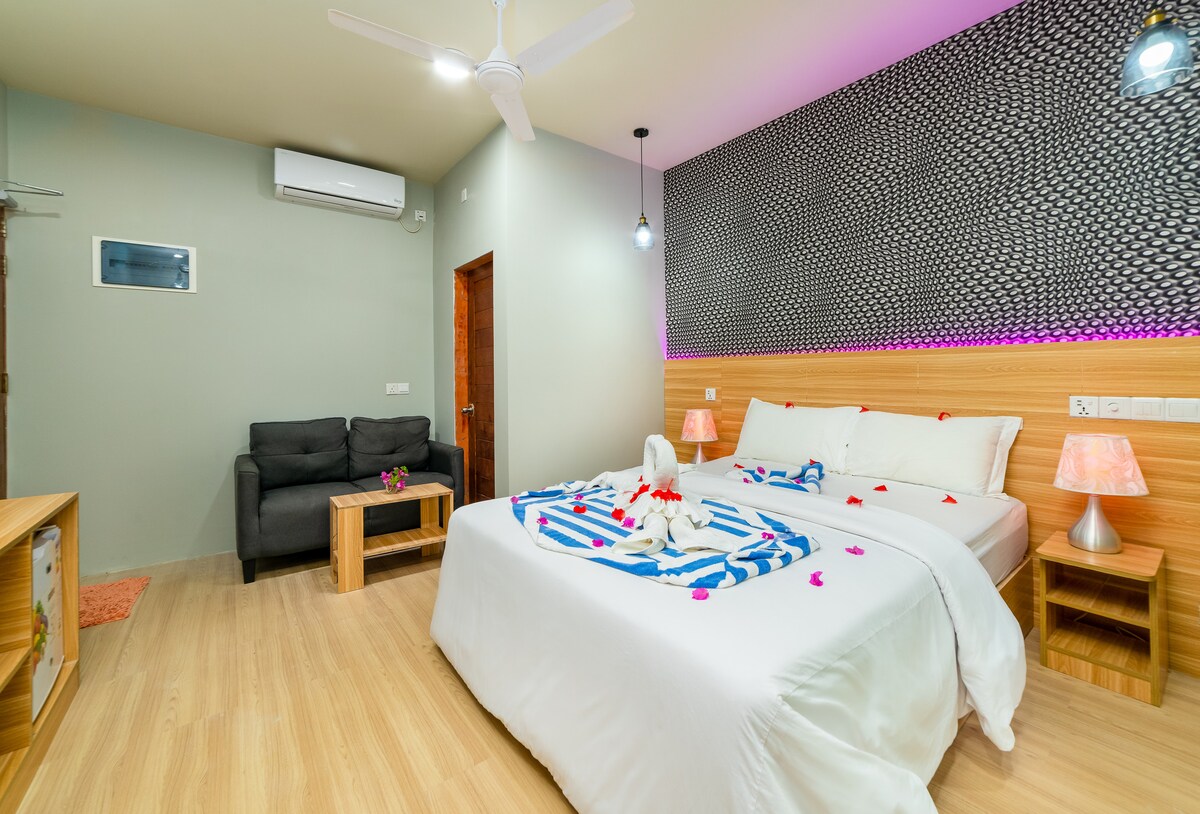 A cozy bedroom features a spacious bed draped with decorative towels and scattered flower petals. A comfortable sofa is positioned against the wall, complemented by a small coffee table. Ambient lighting is provided by bedside lamps and recessed ceiling lights.