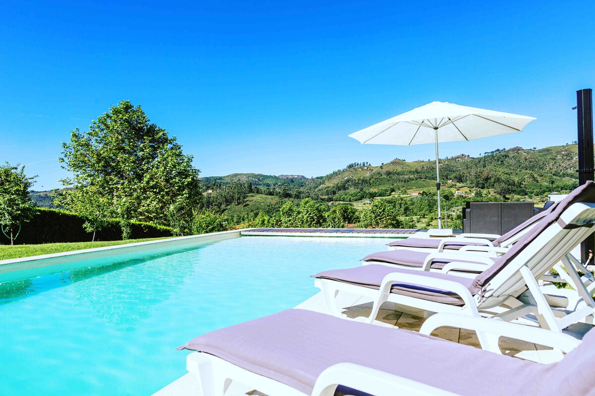 A refreshing swimming pool is set against a backdrop of rolling green hills. Lounge chairs with purple cushions are positioned alongside the pool, while a large white umbrella provides shade. Clear blue skies enhance the tranquil atmosphere.