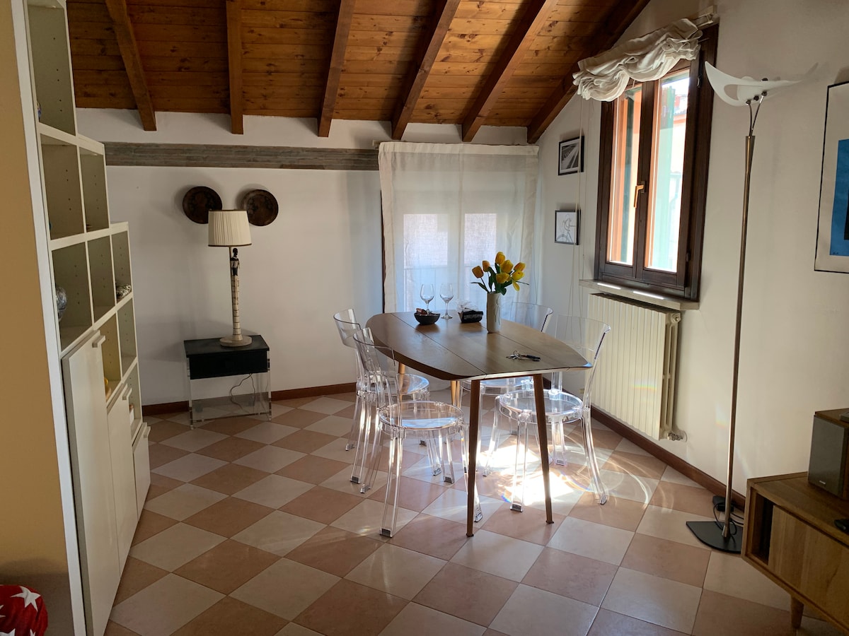 A dining area is situated underneath a wooden beamed ceiling, featuring a round table surrounded by transparent chairs. A floor lamp stands nearby, and natural light filters through the sheer curtains on the window, complementing the tiled flooring.