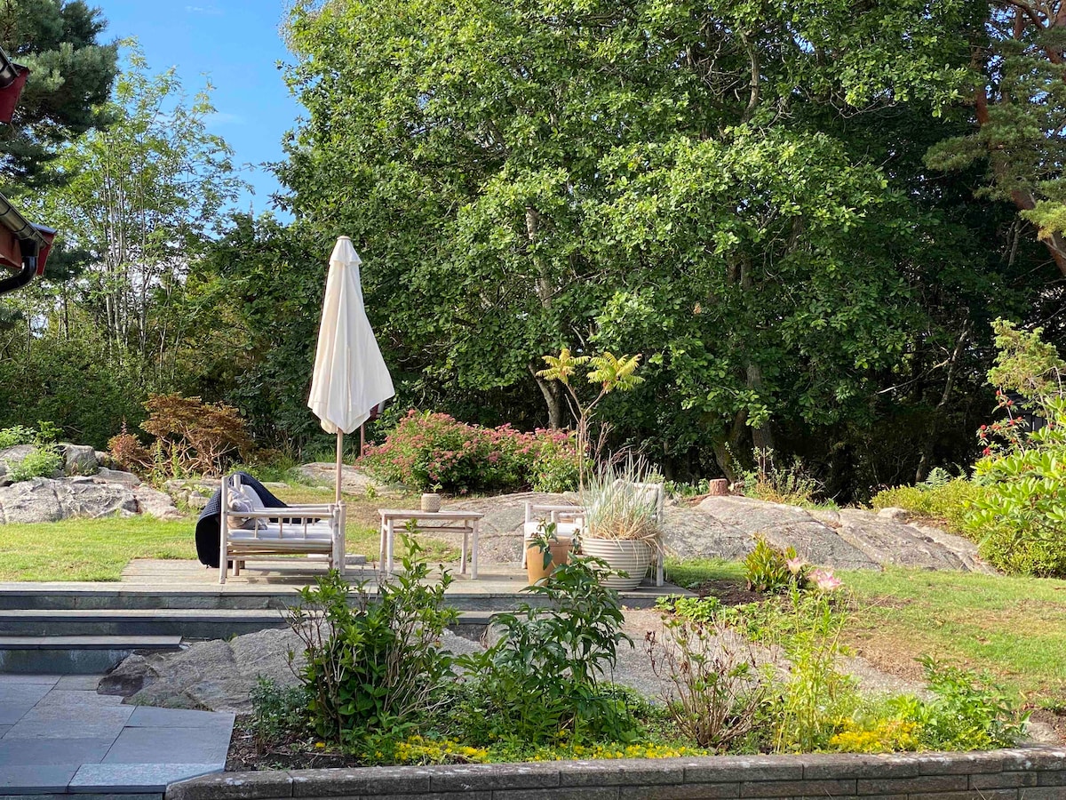 A spacious garden features a lounging area with a sun umbrella and comfortable seating. Surrounding greenery includes decorative plants and shrubs. Natural rock formations are present, enhancing the outdoor space under a clear blue sky.