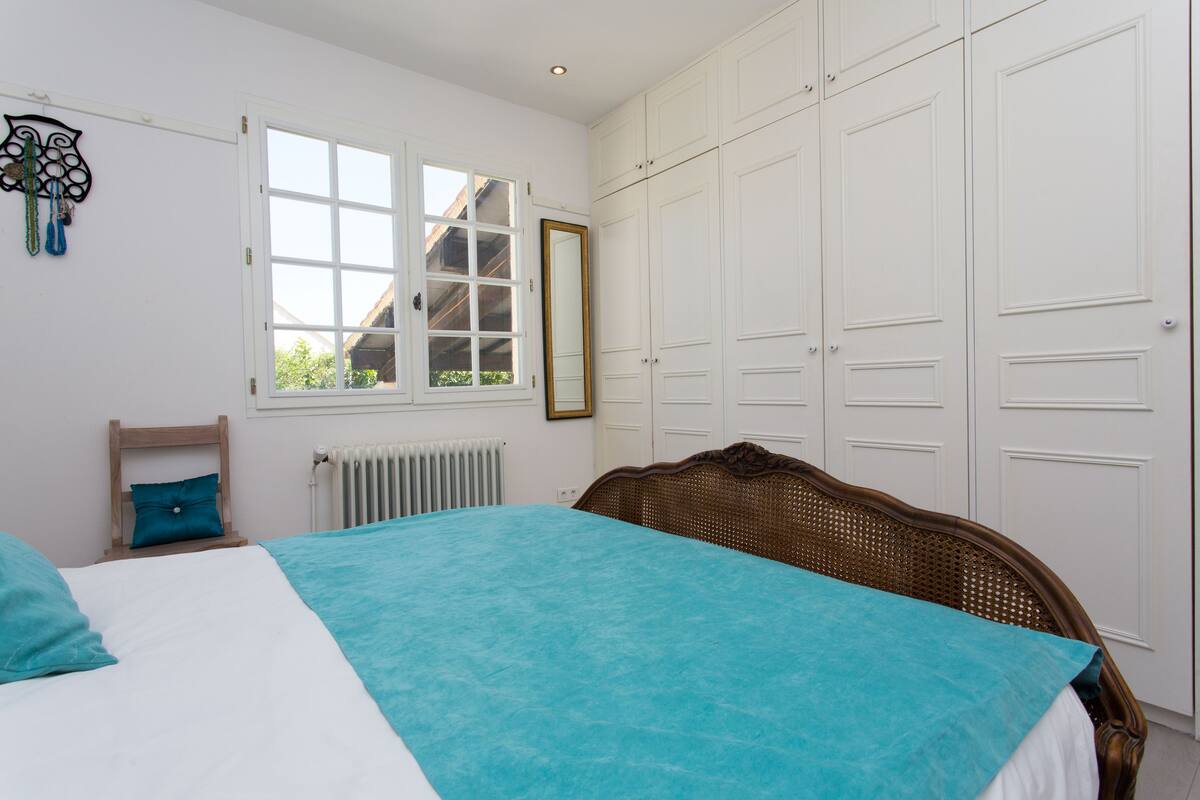A comfortable sleeping area is featured, showcasing a spacious bed with a light blue cover. White cabinetry lines the wall, providing ample storage. Natural light filters in through the multi-pane window, creating a bright and airy environment.
