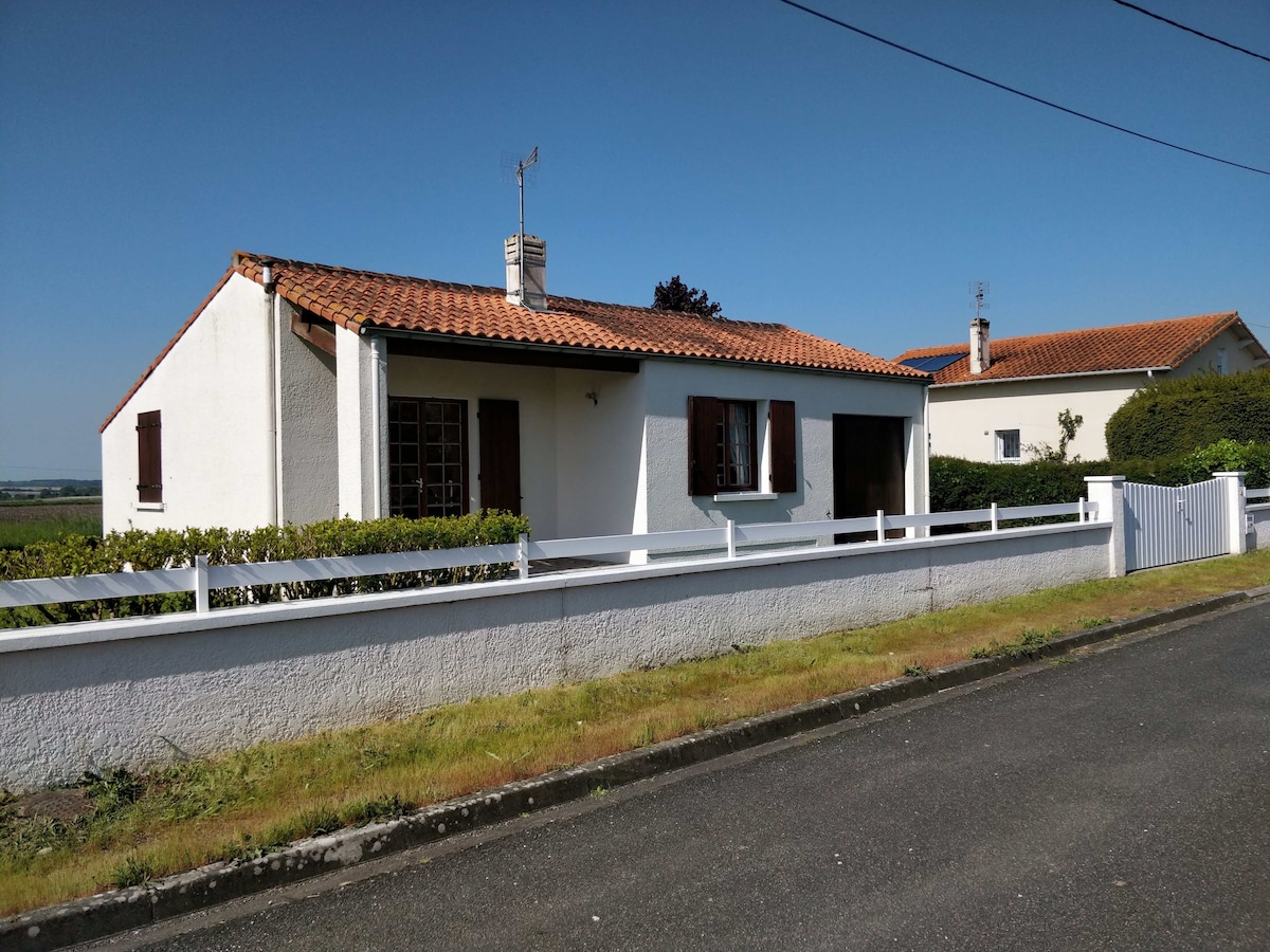 The house is presented with a white exterior and a terracotta roof, featuring several windows covered by wooden shutters. A low white fence borders the property, with neatly trimmed hedges in front, and the surrounding area is characterized by open space.