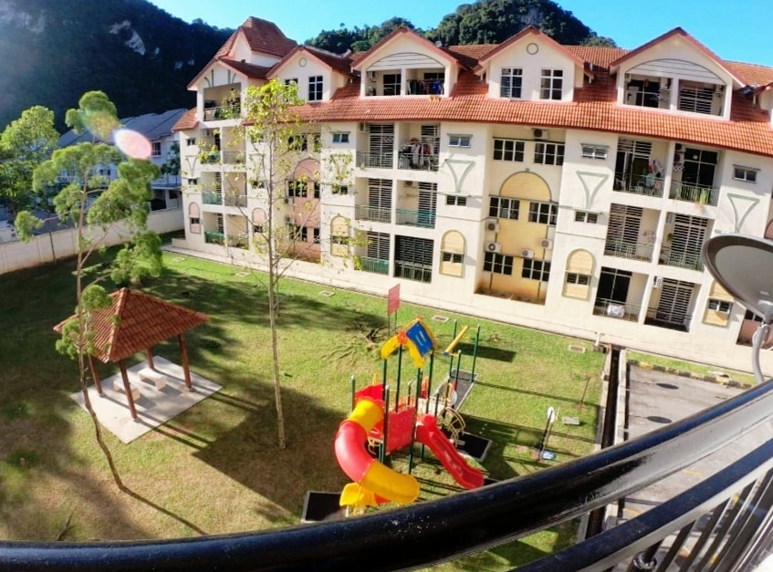 The image showcases the Alpine Village Apartment complex, featuring a playground with a multi-colored slide and swings. A gazebo is visible on the lawn, surrounded by grassy areas and trees. The building's architecture includes large, arched windows and a terracotta roof, set against a backdrop of hills.