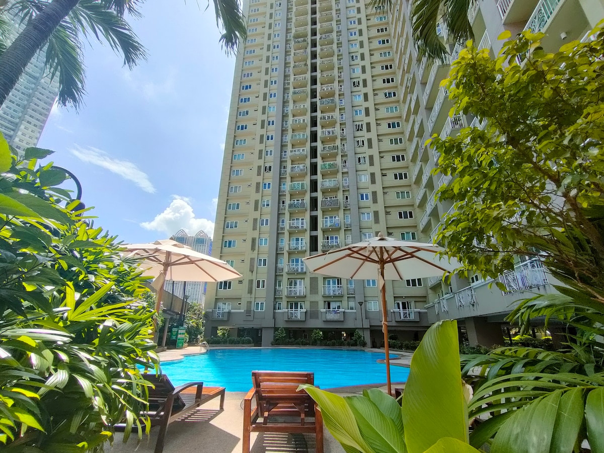 A serene swimming pool is framed by lush greenery and palm trees, with sun loungers positioned under umbrellas. The tall condominium building looms in the background, reflecting a possible urban lifestyle.
