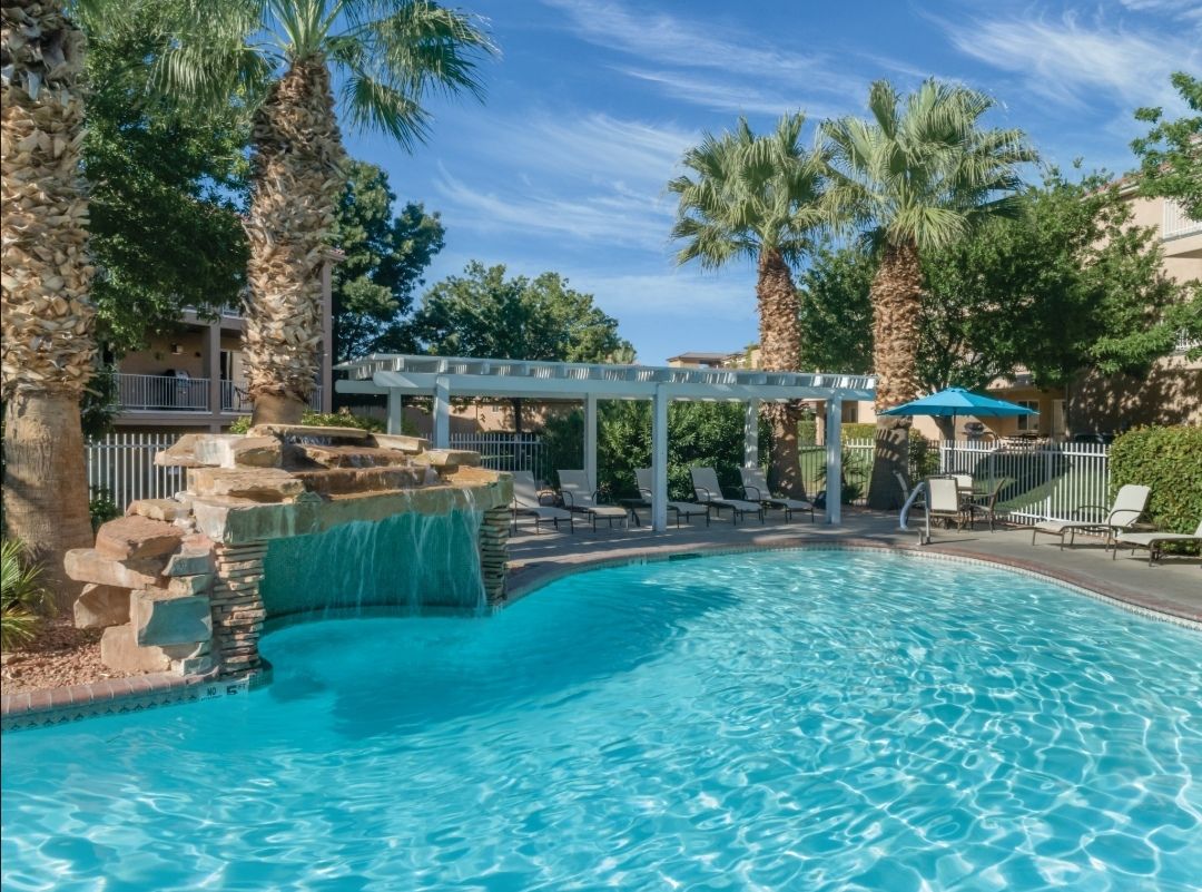 A serene outdoor pool area is highlighted by a waterfall feature and surrounded by palm trees. Comfortable lounge chairs are positioned around the pool, which is filled with clear blue water, offering a refreshing retreat in a peaceful setting.