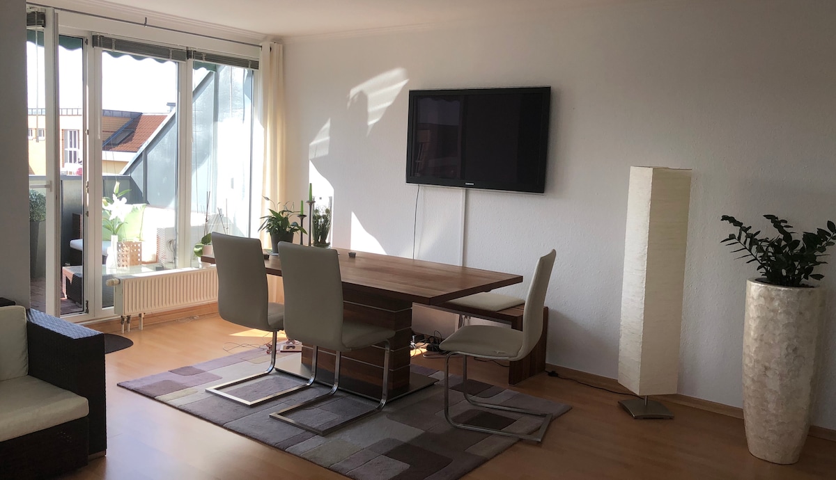 A bright living area is presented with a dining table and four chairs. A flat-screen television is mounted on the wall, and sunlight illuminates the space. Indoor plants are positioned by the large glass doors leading to a balcony, enhancing the inviting environment.
