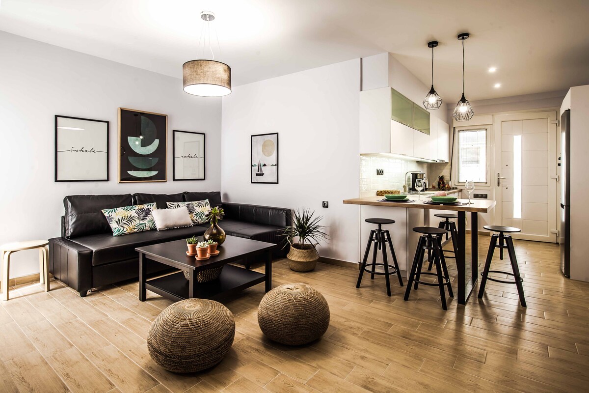 The living area showcases a sleek black sofa positioned across from a dark coffee table, complemented by seating at a kitchen island. Unique wall art enhances the modern aesthetic, while natural light fills the space through a nearby door. Earth-toned textures are present throughout the room.