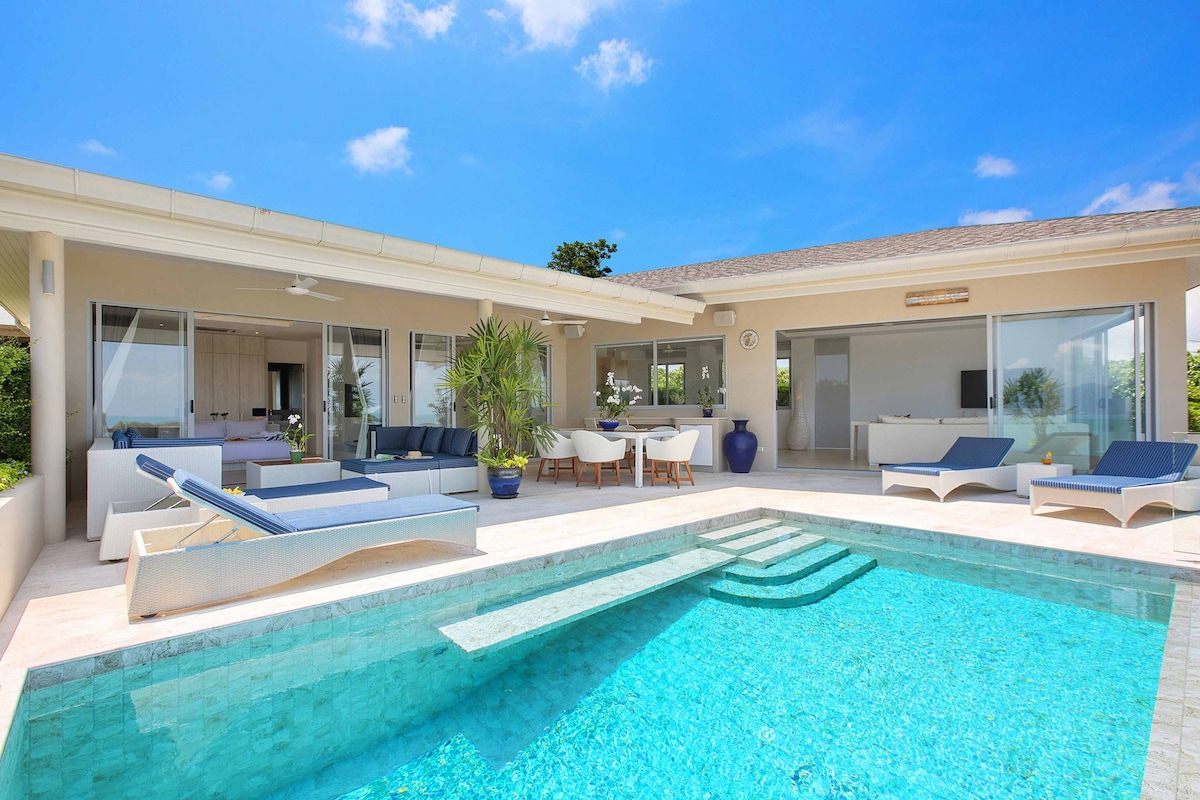 A modern villa exterior showcases a spacious outdoor area with an inviting infinity pool. Relaxing lounge chairs are arranged nearby, alongside a dining area under a covered patio. Large glass doors create a seamless connection to the interior living spaces, accentuated by blue skies and lush greenery.