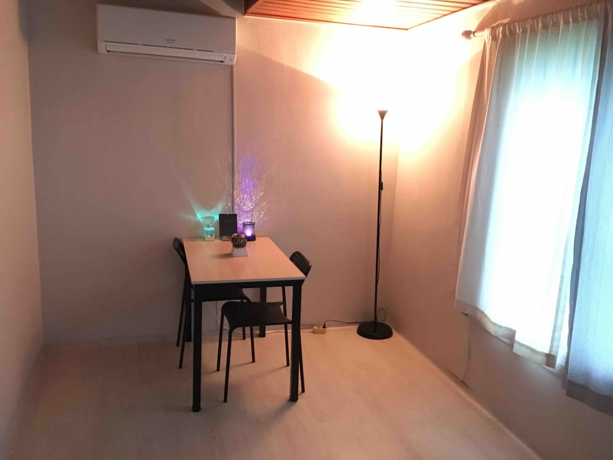 A simple dining area features a wooden table surrounded by black chairs. Soft light from a nearby floor lamp illuminates the space, complemented by natural light filtering through a sheer curtain. An air conditioning unit is mounted above, ensuring comfort.
