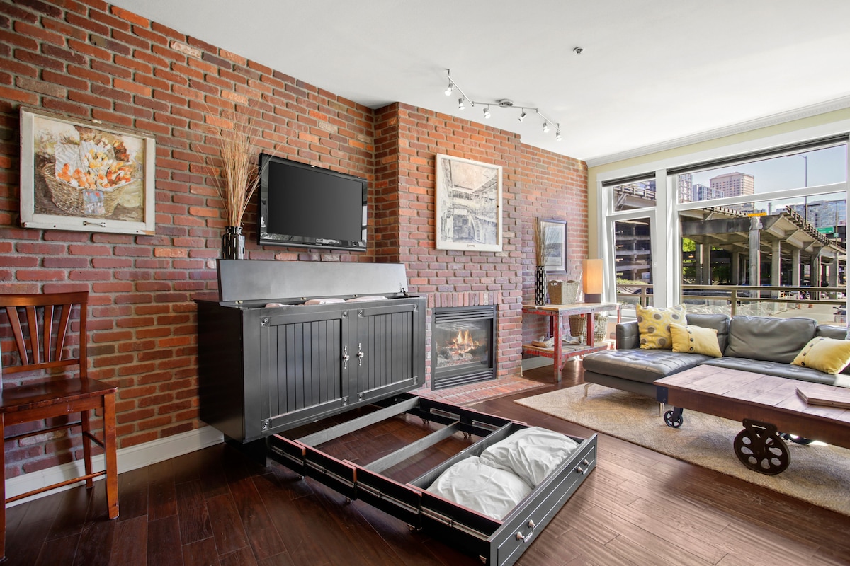 A sunlit living room features red-brick accent walls and espresso wood floors. A comfortable leather couch and chair are arranged near a gas fireplace, with a TV mounted above a stylish cabinet. Large windows provide views of the outdoor space.