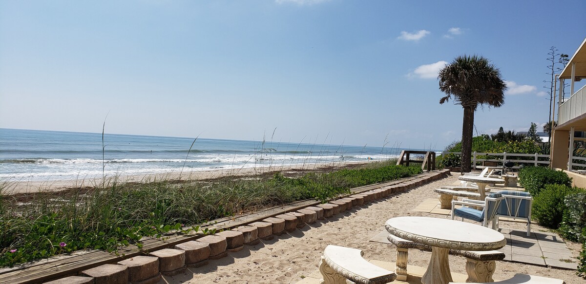 A sandy area with stone tables is shown alongside the coastline, where gentle waves meet the shore. Lush greenery borders the beach, while a palm tree provides shade. The clear blue sky enhances the tranquil atmosphere of the beachfront, overlooking the ocean.