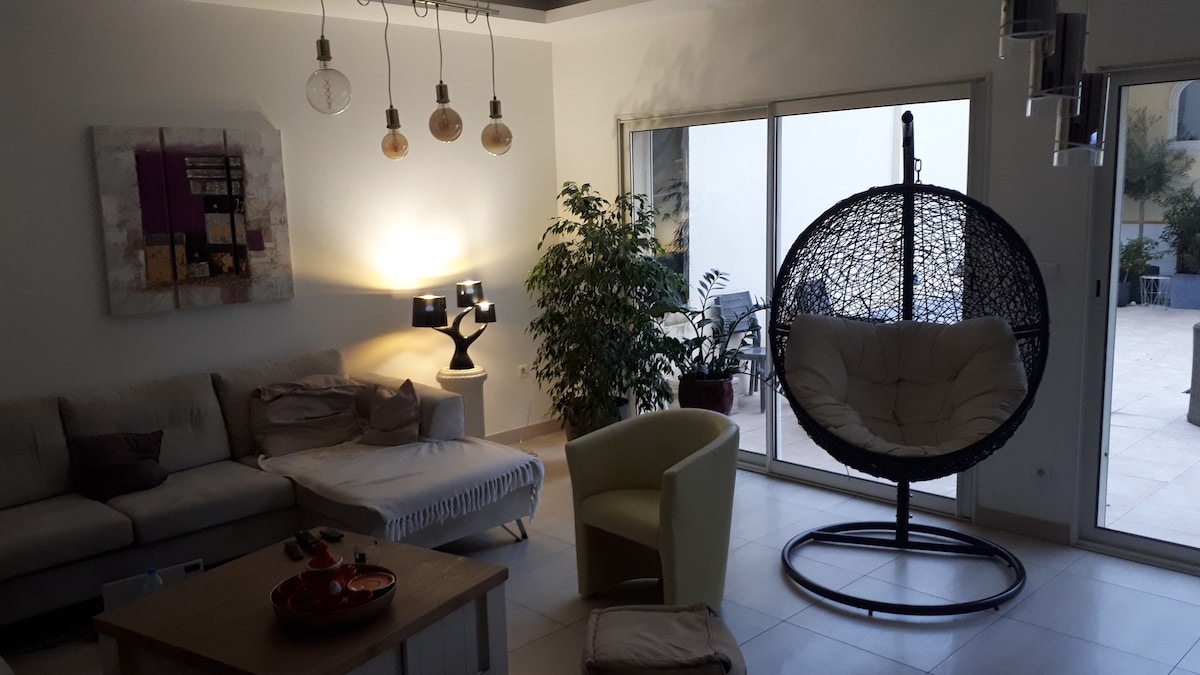 A spacious living area features a comfortable sofa and a unique hanging chair. Decorative lamps provide soft lighting, while a large window invites natural light and views of the outdoor patio. A potted plant adds a touch of greenery to the tranquil ambiance.