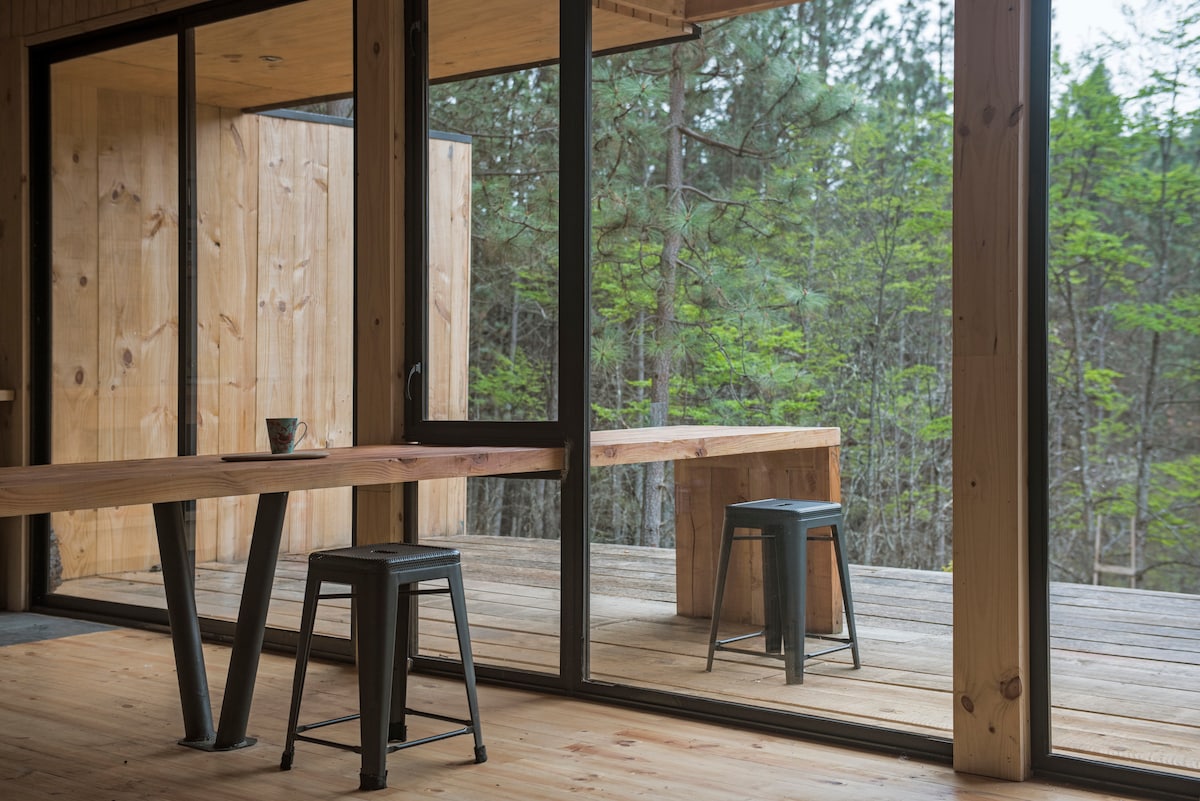 A spacious dining area features a wooden table elegantly paired with metal stools. Large glass doors provide a view of the outdoor deck surrounded by greenery, creating a connection between indoor and outdoor spaces. Natural light fills the room, enhancing the warm wood tones.
