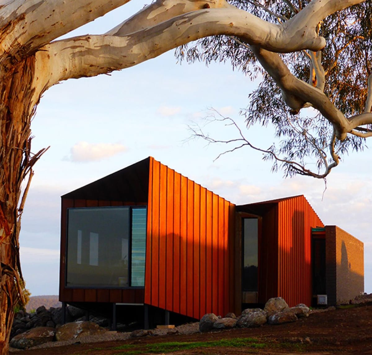 Una casa tipo contenedor hecha de metal naranja se destaca contra un cielo abierto, con un árbol de eucalipto en primer plano.