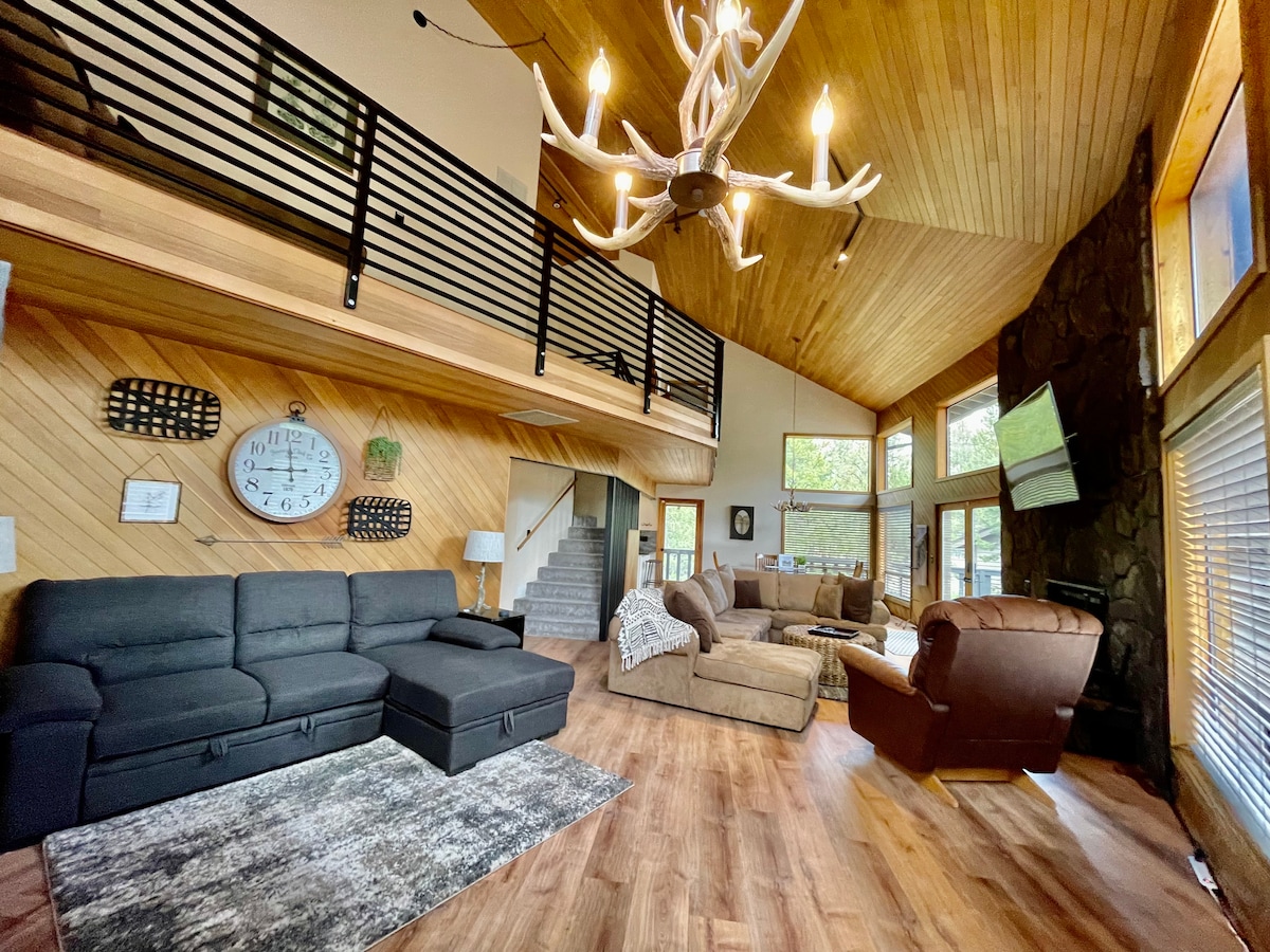 A spacious living room features vaulted wooden ceilings and large windows, allowing natural light to fill the area. Various seating options include a sectional sofa, a plush armchair, and a comfortable couch. Decorative elements such as a large clock and a unique light fixture add character to the space.