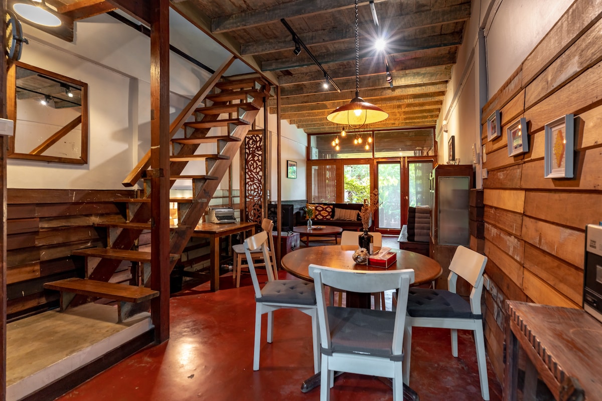 The living and dining area features a round table surrounded by four chairs, with wooden accents throughout. A staircase leads to the second floor, illuminated by soft lighting. Large glass doors provide a view into a lush outdoor space, enhancing the inviting atmosphere.