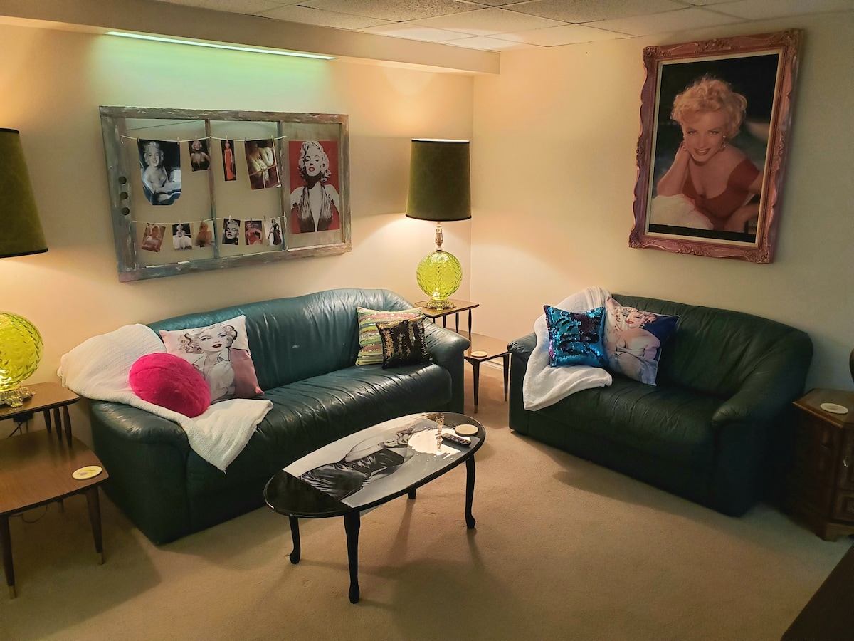 A cozy living area features two leather sofas adorned with colorful pillows. A coffee table sits in the center, displaying decorative items. Warm lamp light creates a relaxed ambiance, complemented by framed art on the walls, adding character to the space.