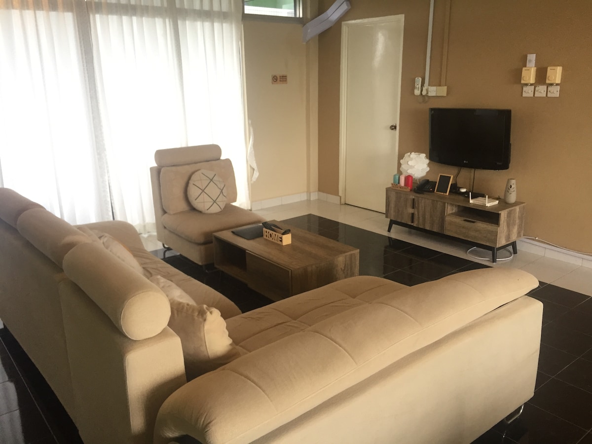A modern living area is presented with a light-colored sectional sofa and a matching armchair. A low wooden coffee table sits on a dark tiled floor, with a television mounted on the wall opposite. Sheer white curtains allow natural light to filter in.