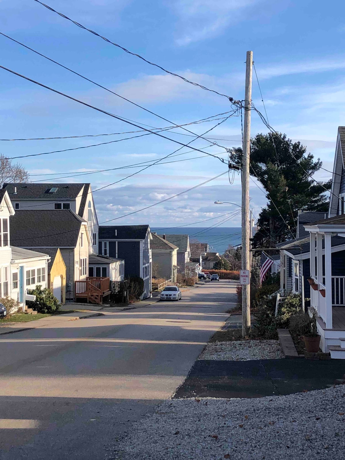 A serene street lined with charming houses leads toward the horizon, where the ocean can be glimpsed at the end. Power lines traverse the scene, and the ground is a mixture of gravel and pavement, creating a welcoming coastal neighborhood atmosphere.