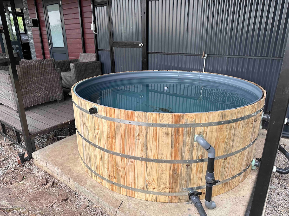 A wooden hot tub is positioned outside on a stone surface, surrounded by a deck. The tub features a cylindrical shape with a smooth, dark interior. Nearby, seating arrangements made from woven material can be seen, offering a space for relaxation.