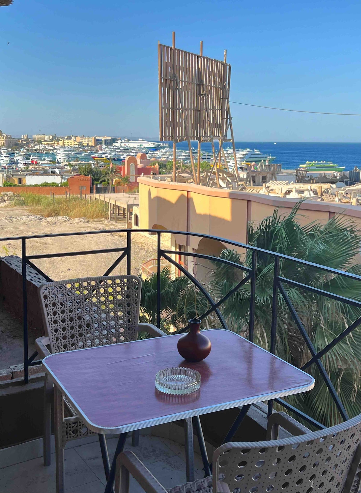 A balcony view showcases a sparkling sea and yachts in the distance, framed by palm trees. A small table with a vase and ashtray is centered on the balcony, inviting relaxation while enjoying the surrounding scenery. A large structure is visible nearby, highlighting the vibrant locale.